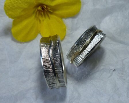 Trauringe Variable Trauringe „Variable“. Ringschiene mit Furchen. 1 Ring mit umlaufendem Goldring und 1 Ring mit umlaufendem Silberring. Breite jeweils 8 mm.